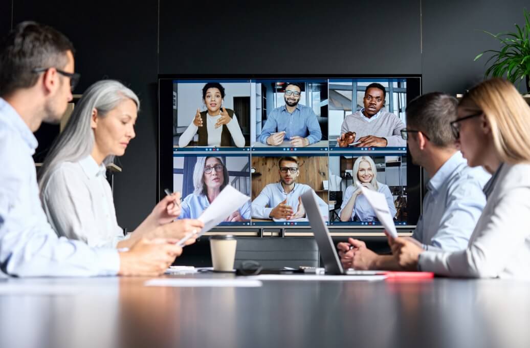 Hybrides Meeting: Teilnehmende am Konferenztisch und per Videokonferenz auf einem großen Bildschirm
